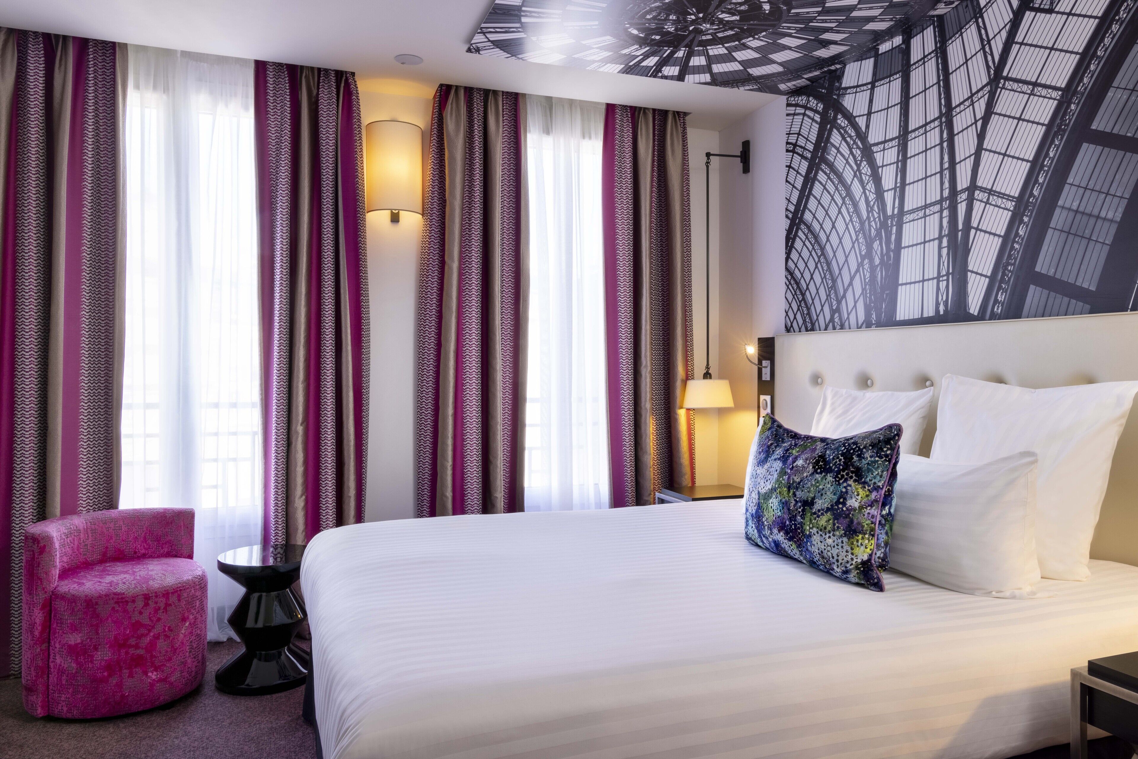 A hotel room with a large bed, a pink armchair, a small round table, and a wall-mounted lamp.