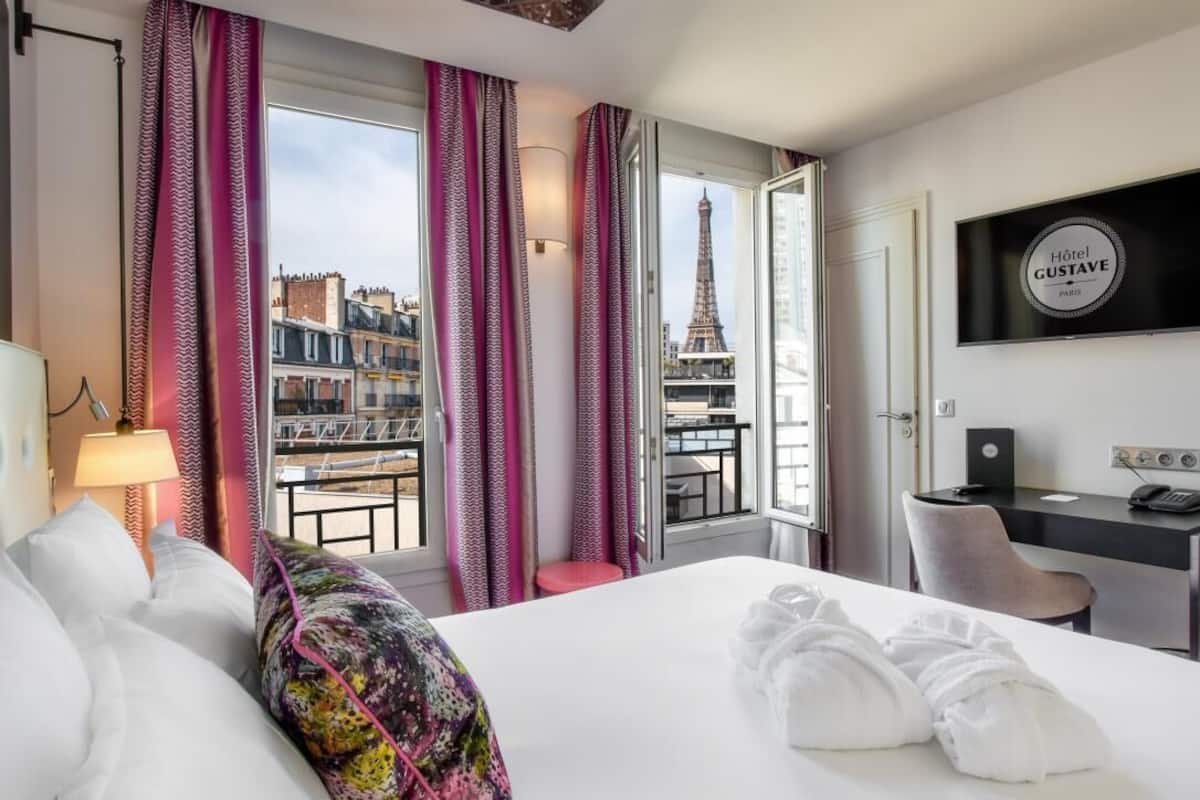 A hotel room with a bed, a desk, a chair, a TV, and a view of the Eiffel Tower.