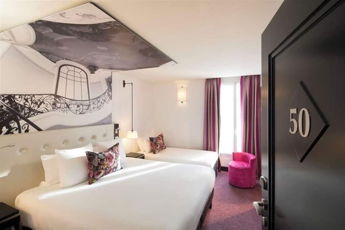 A hotel room with two beds, a large painting on the wall, a pink chair, and a door with the number 50.