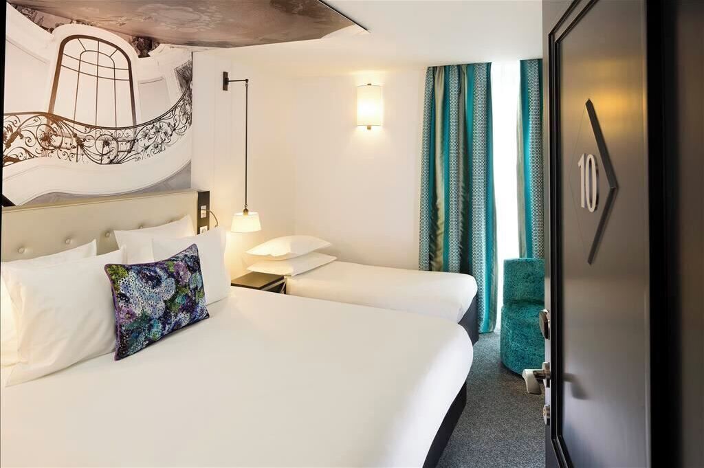A hotel room with a large bed, a smaller bed, a nightstand, a lamp, a chair, and a door with a number.