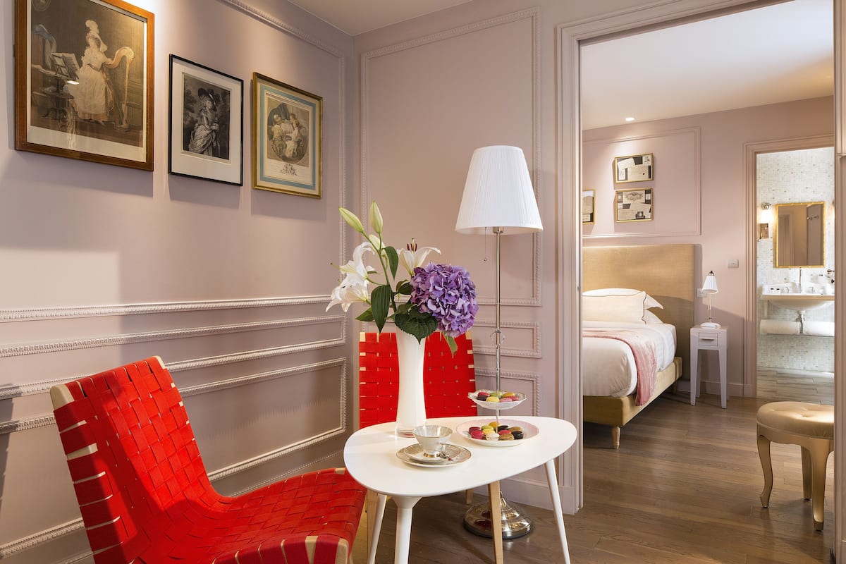 Habitación de hotel con cama, mesita de noche con lámpara, pequeña mesa con un florero y una silla roja.