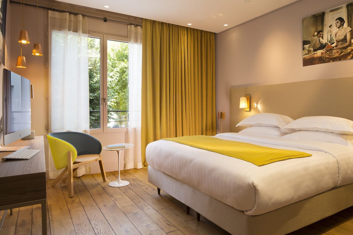 Una habitación de hotel con cama, escritorio, computadora, silla y ventana con cortinas.