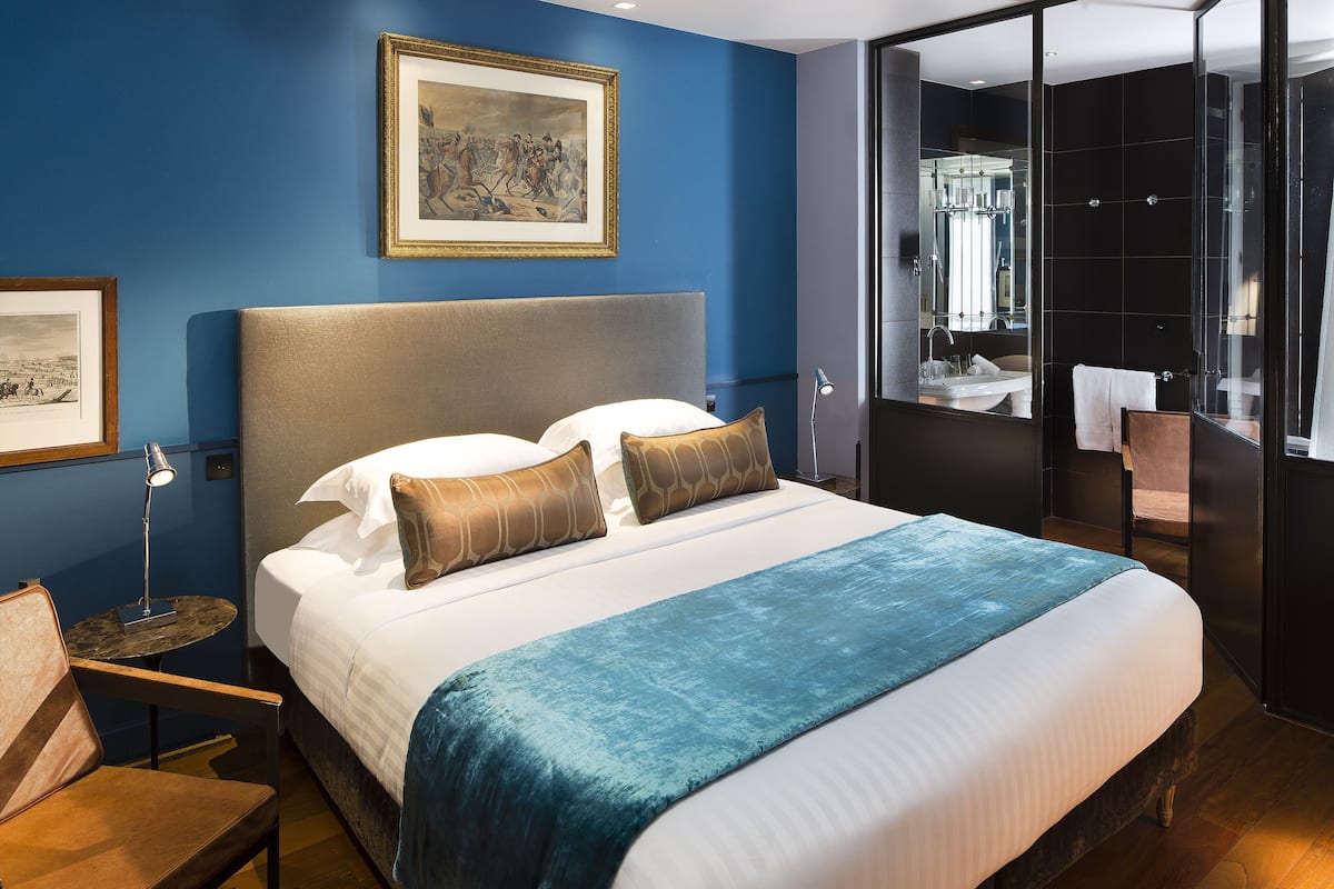 Habitación de hotel con cama, mesita de noche, una silla, un cuadro en la pared y un baño visible a través de una puerta abierta.