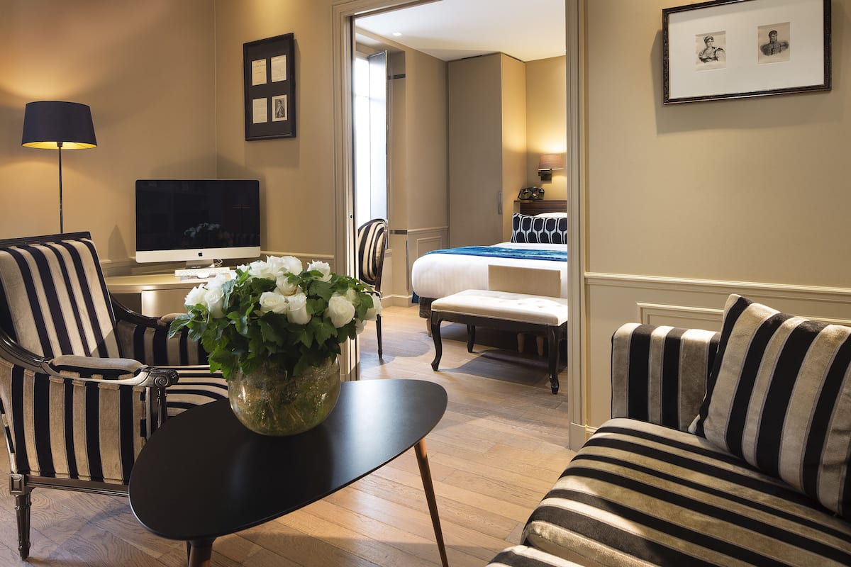 Habitación de hotel con sofá a rayas, una mesa negra con un florero y un televisor de pantalla plana, cama con cubrecama a rayas y un cuadro en la pared.
