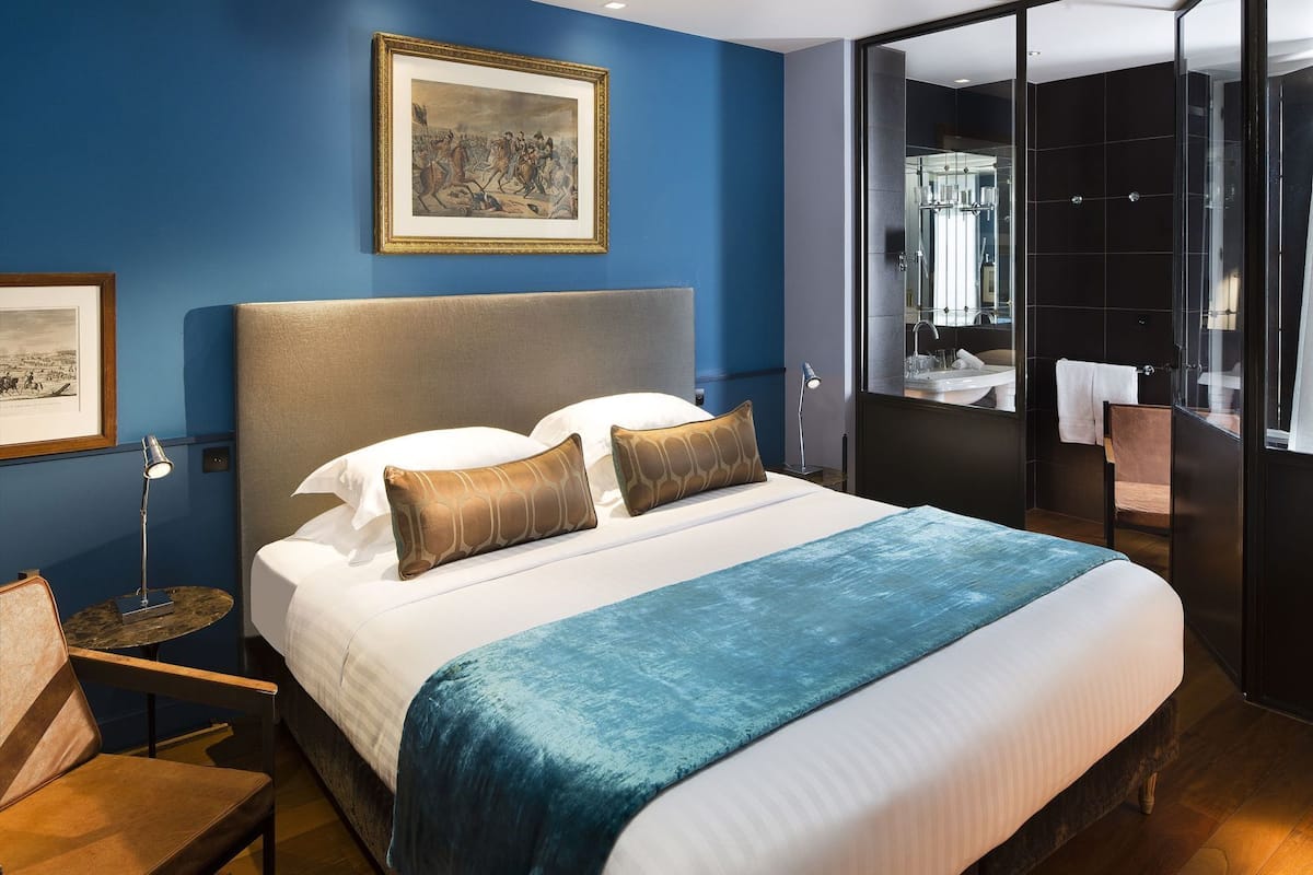 Una habitación de hotel con una cama grande, un cuadro en la pared, un baño con puerta de cristal y una silla.