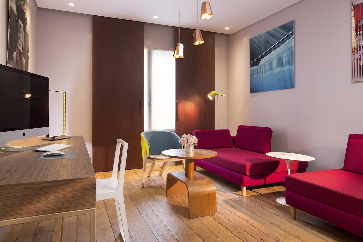 Una habitación moderna con un escritorio de madera, una computadora, un televisor de pantalla plana, un sofá rojo, una silla azul y una pequeña mesa redonda con flores.
