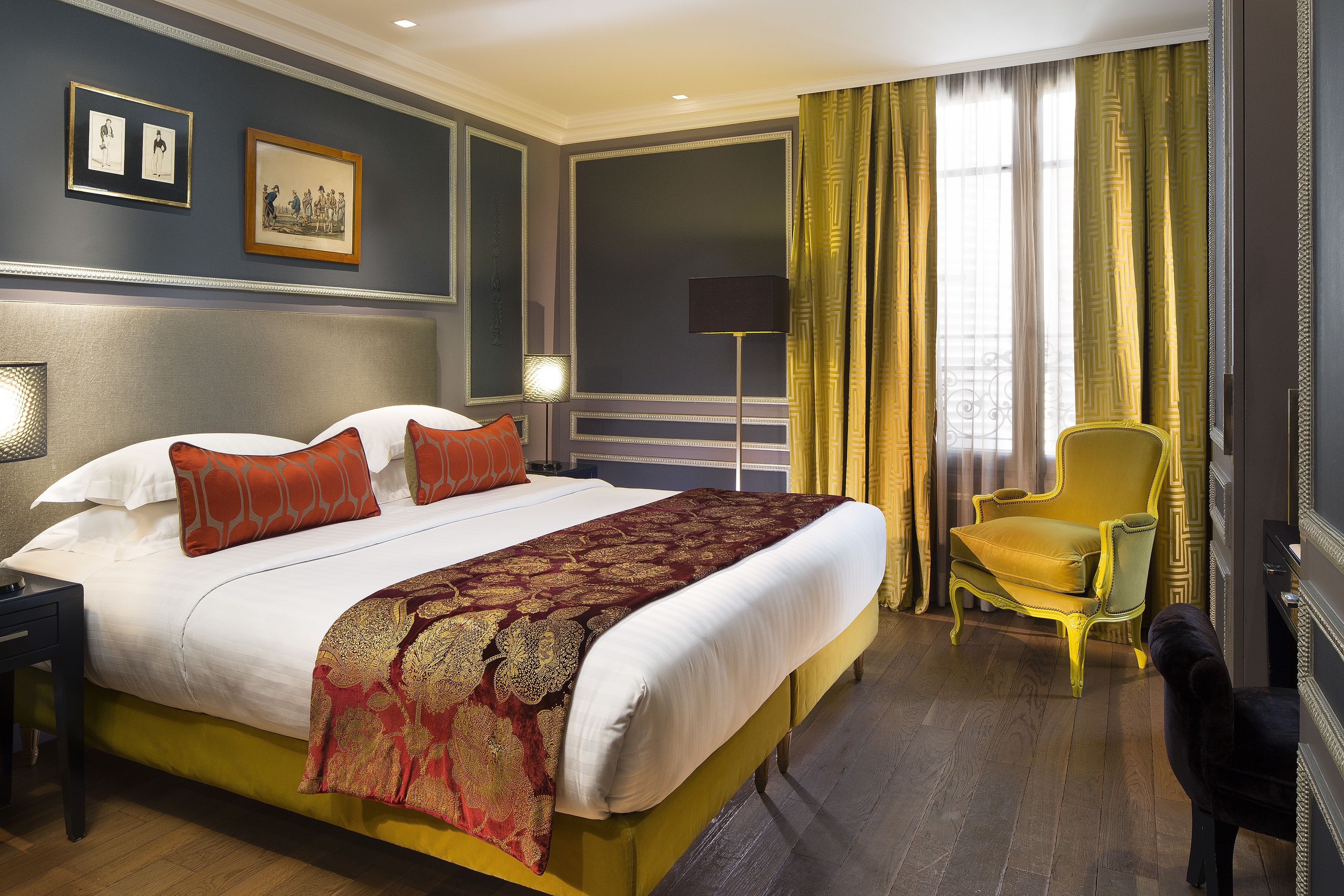 Una cama grande con colcha blanca y roja, un sillón amarillo y una silla negra en una habitación con paredes oscuras y suelo de madera.