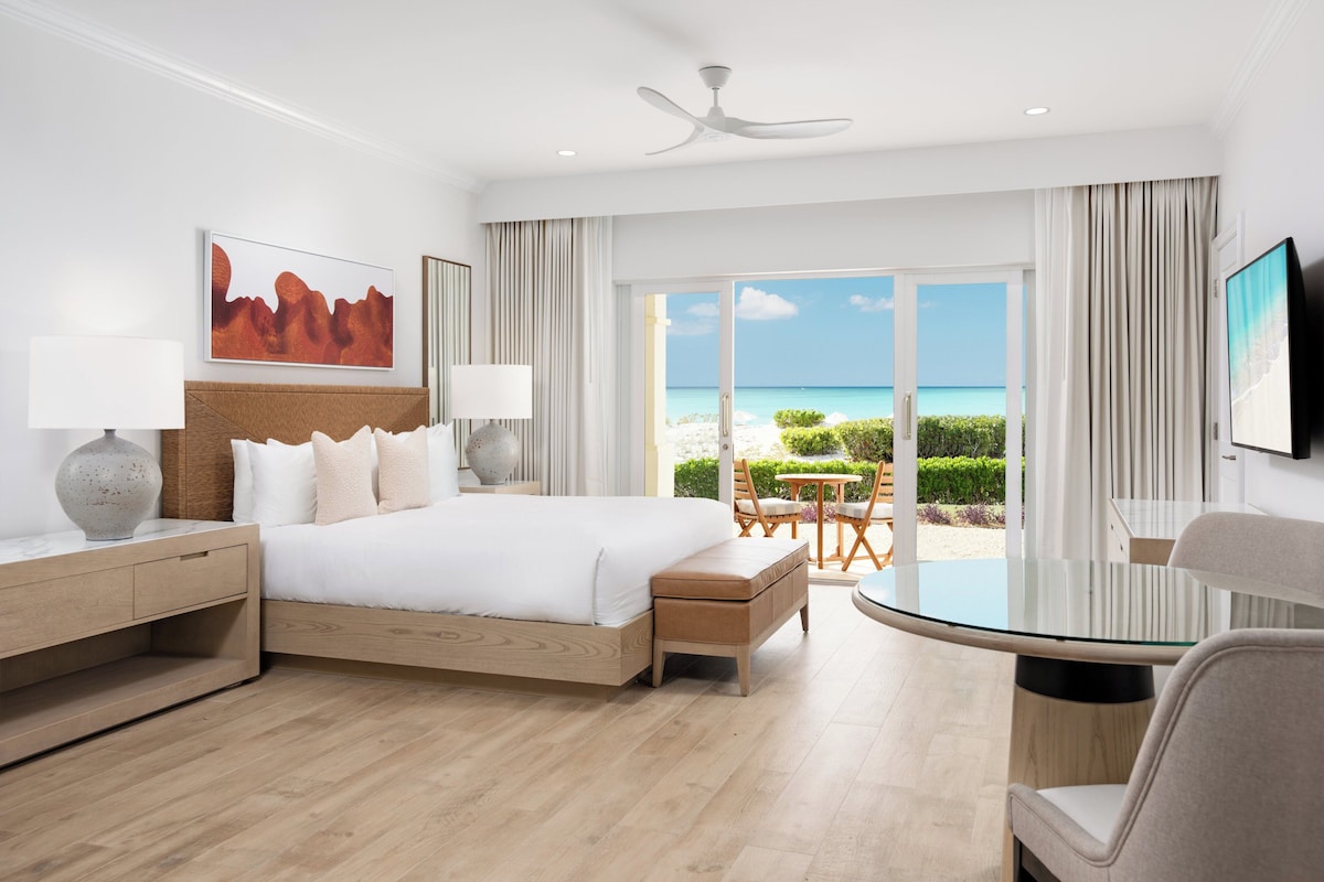 A modern hotel room with a large bed, a glass table, and a view of the beach.