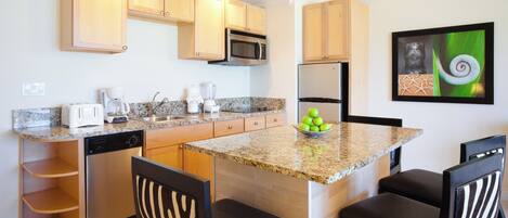 A modern kitchen with granite countertops, wooden cabinets, a microwave, a coffee maker, and a bowl of green apples on the island.