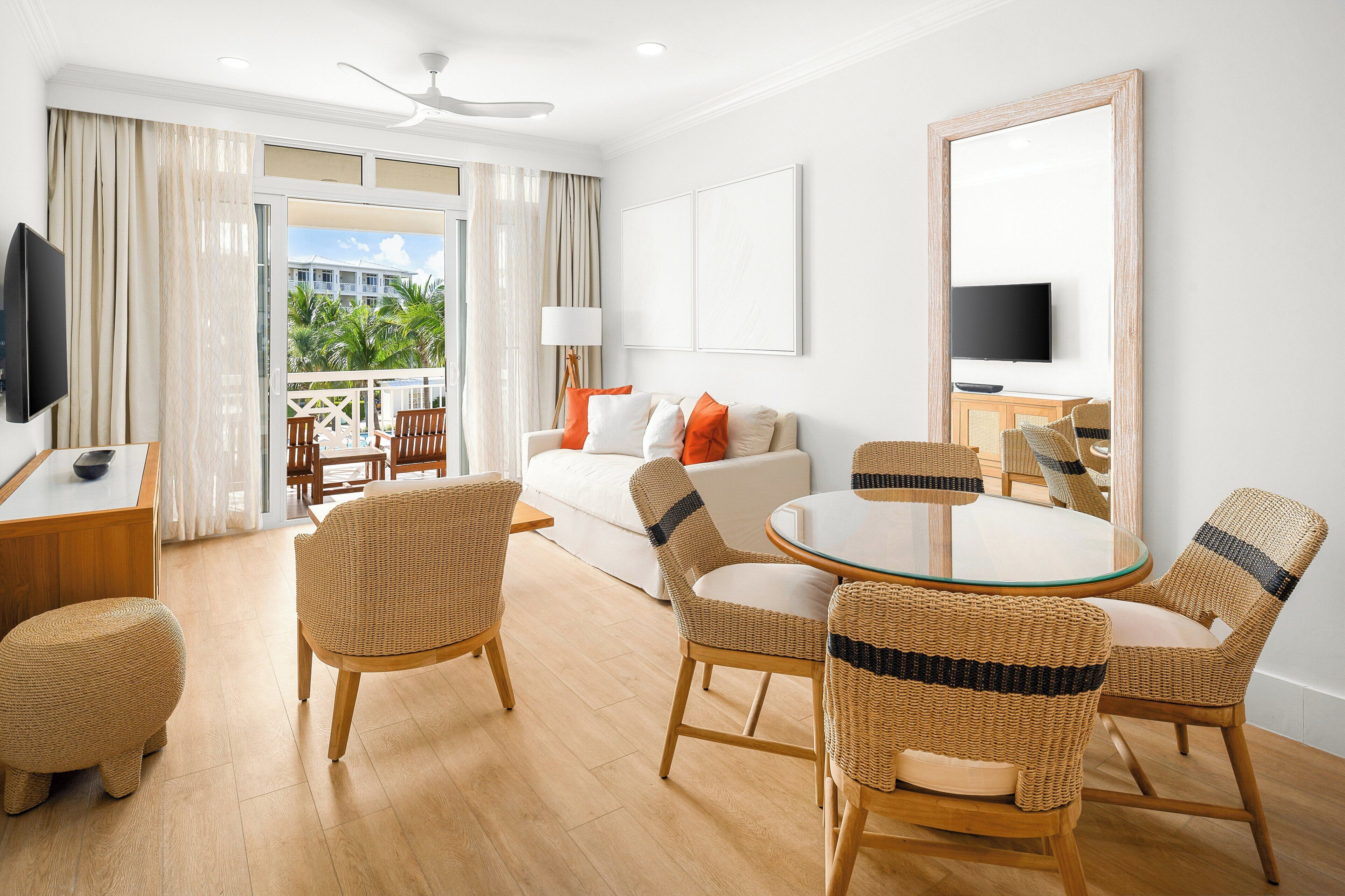 Habitación de hotel con balcón, mobiliario de mimbre, una mesa redonda de vidrio y un televisor en la pared.