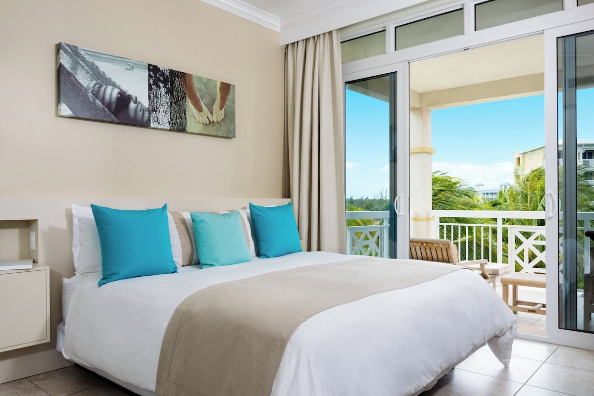 A hotel room with a bed, a painting, a balcony with a view, and a chair.