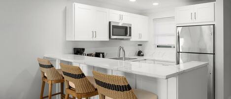 A kitchen with white cabinetry, a stainless steel refrigerator, a microwave, and a sink. There are three wicker bar stools with cushions.