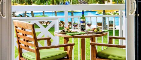 A wooden chair with a green cushion, a wooden table with a bottle and glasses, and a view of a pool and palm trees.
