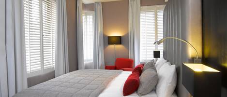 A modern hotel room with a large bed, a red armchair, and a desk with a lamp.
