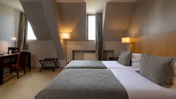 Una camera d'albergo con due letti, una scrivania e una sedia. Due finestre con tende, una lampada e un termosifone.