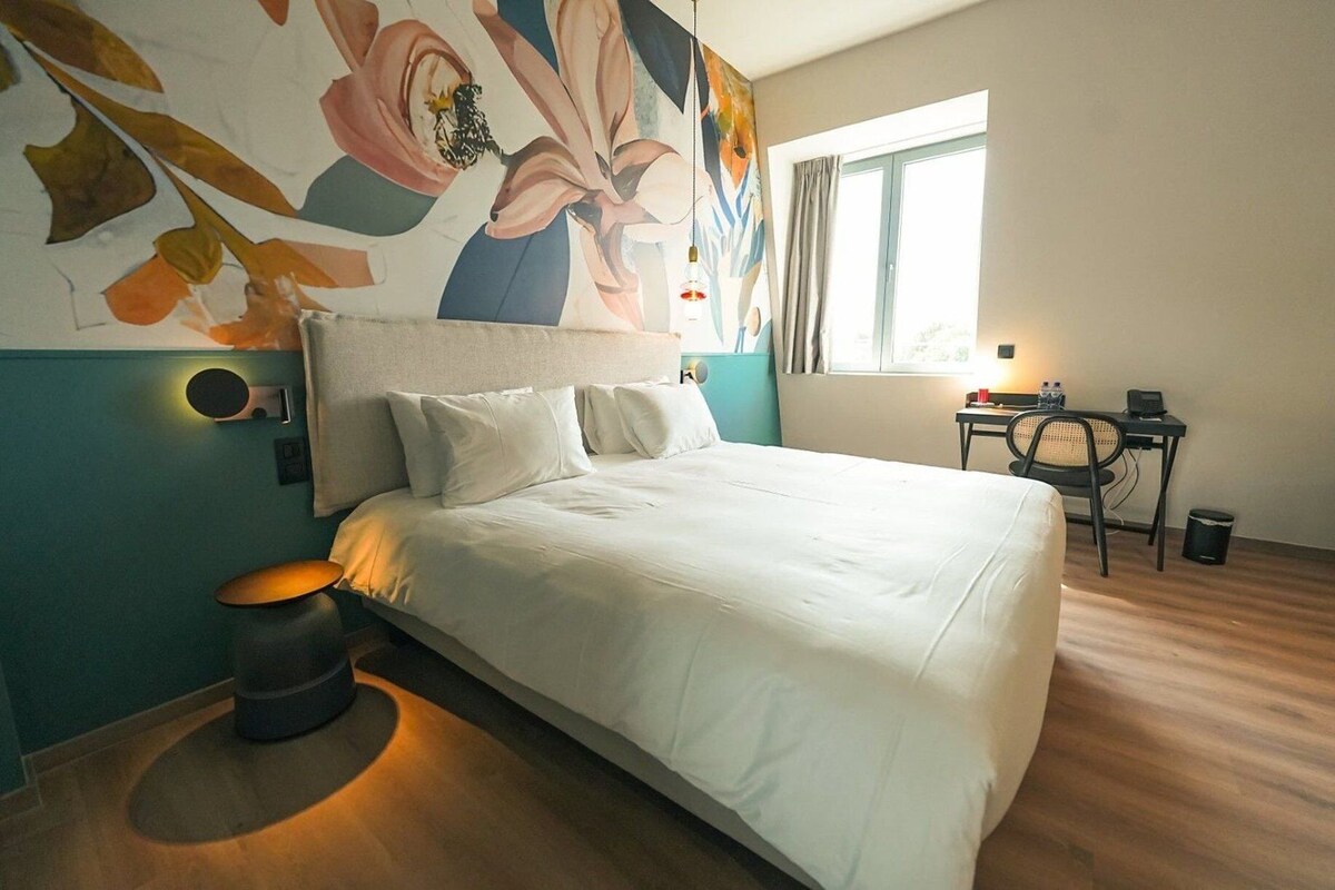 Quarto de hotel com uma cama grande, uma mesa de trabalho, uma cadeira e uma janela com cortinas.