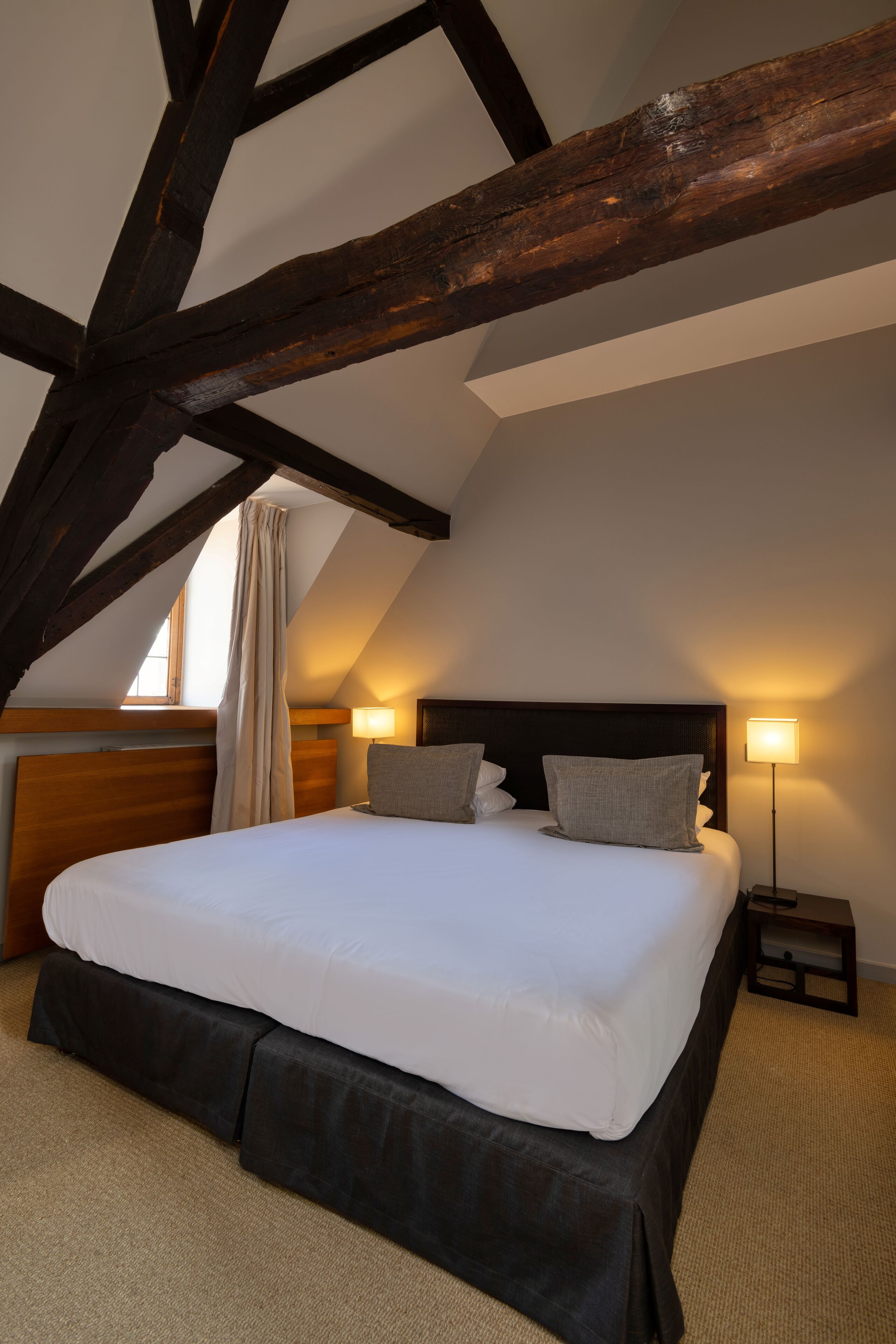 A bedroom with a large bed, wooden beams, a window with curtains, and two bedside lamps.