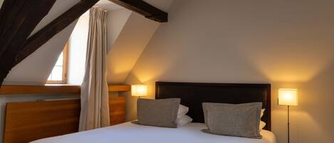A bedroom with a large bed, wooden beams, a window with curtains, and two bedside lamps.