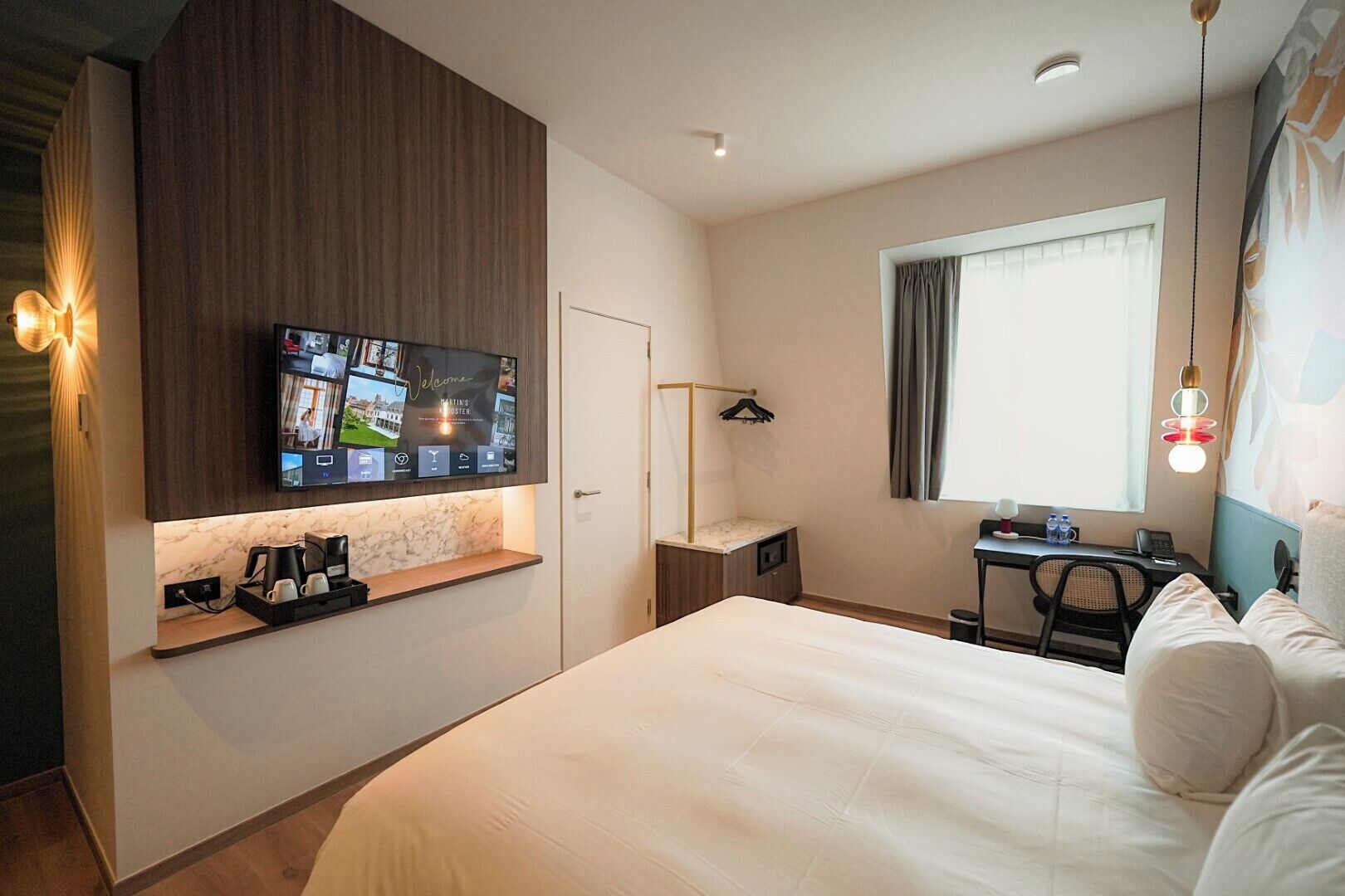 A modern hotel room with a large bed, a desk with a computer, a TV mounted on the wall, and a window with curtains.