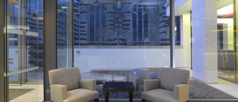 A modern interior with two armchairs and a small table, a large window with a view of a historic building, and a glass partition.