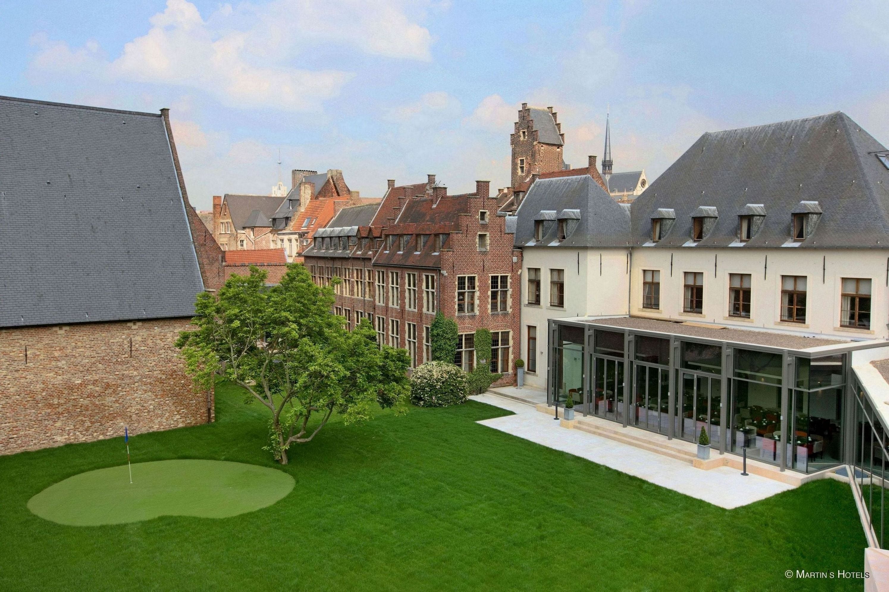 A courtyard with a green lawn, a modern glass building, and traditional brick buildings in the background.