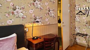 A hotel room with floral wallpaper, a bed, a desk with a lamp, and a chair.