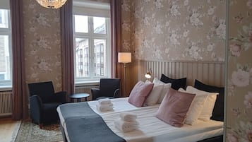Quarto de hotel com uma cama, uma cadeira, uma mesa, um abajur e vista para edifícios pela janela.