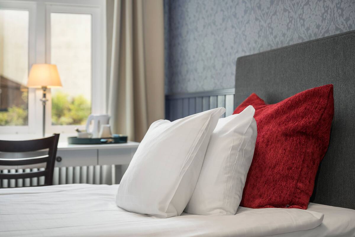 Cama bem arrumada com almofadas brancas e vermelhas, cabeceira cinza e janela com abajur ao fundo.