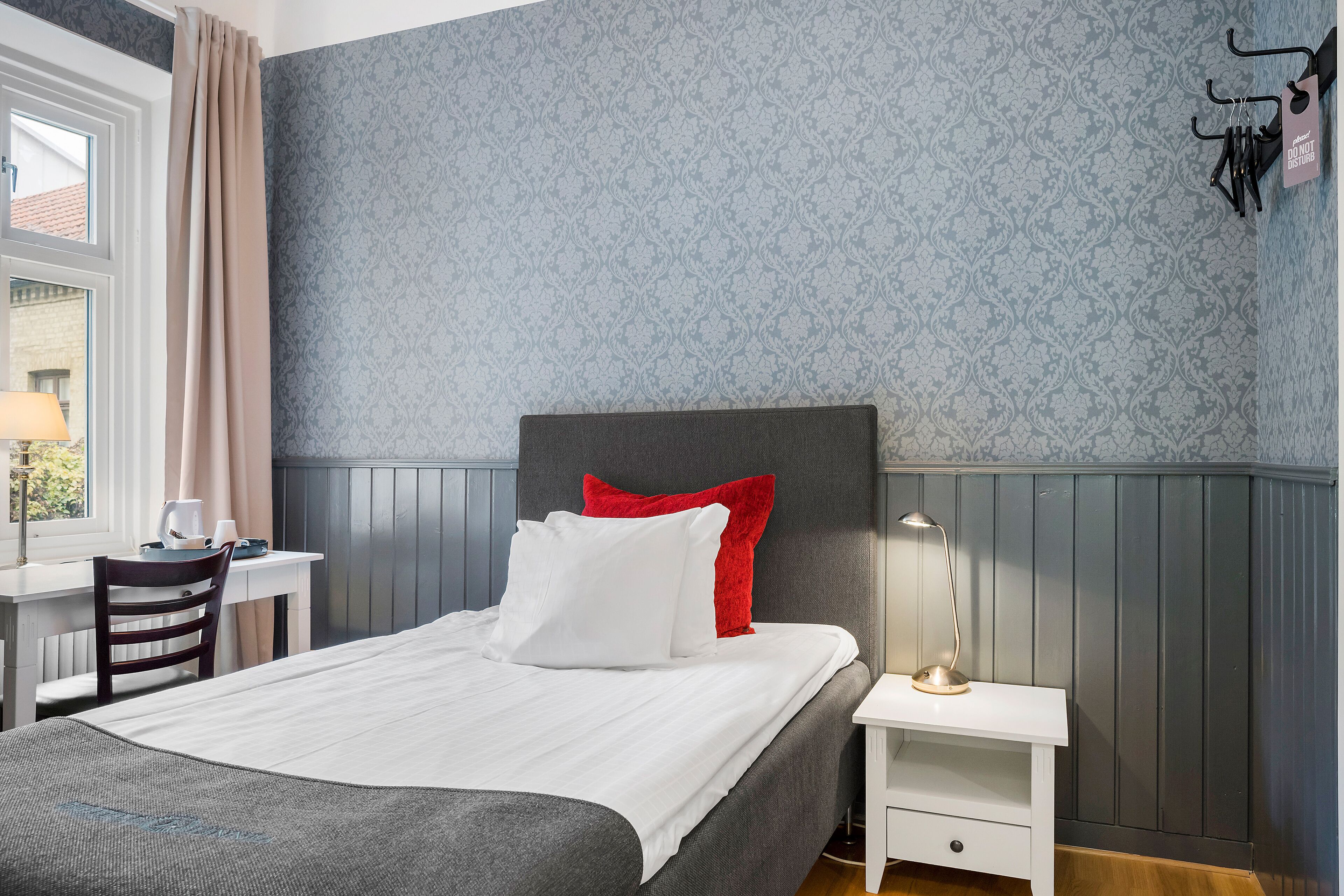 A neatly made bed with white and red pillows, a bedside table with a lamp, and a chair.