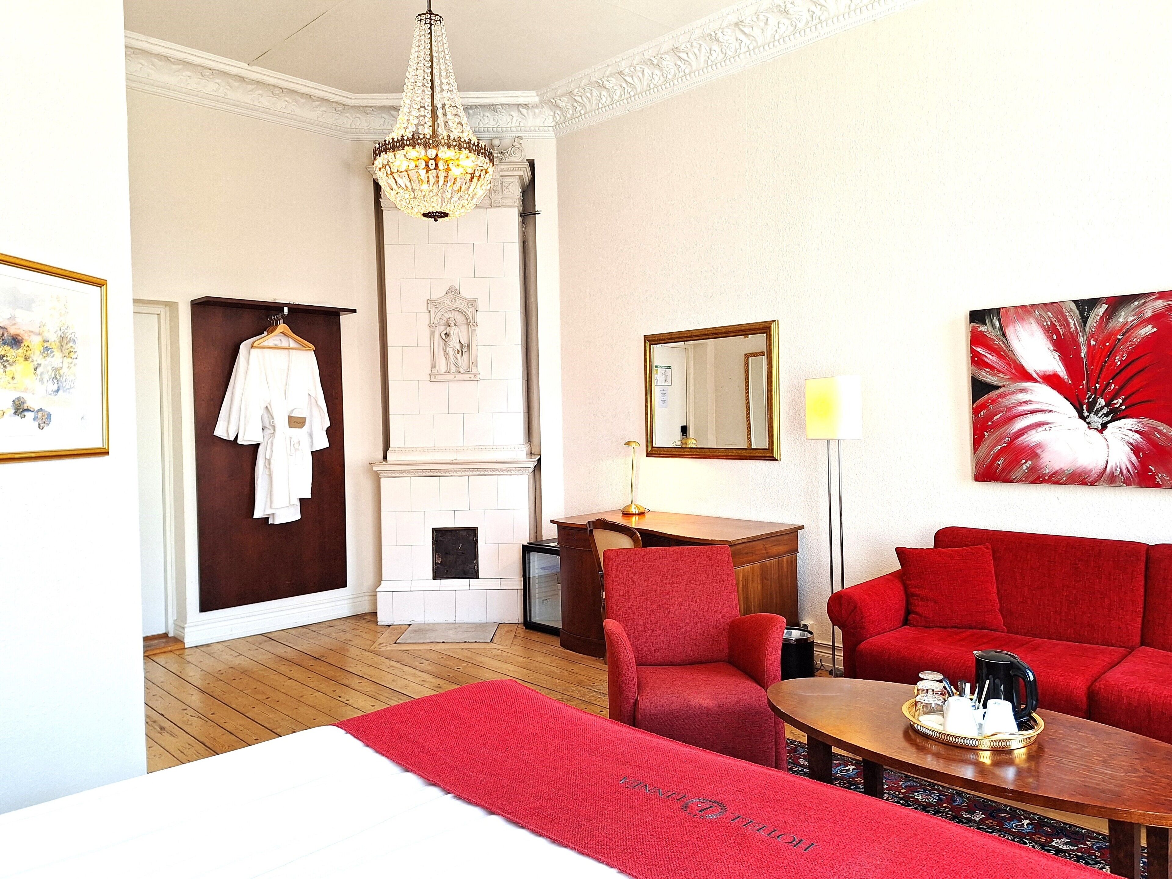 A hotel room with a red sofa, a wooden desk with a tray of items, a red chair, a white bed, a chandelier, a fireplace, and a painting.
