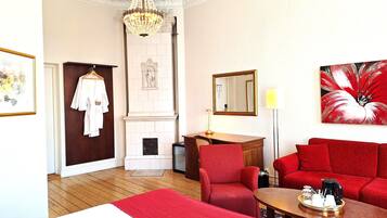 A hotel room with a red sofa, a wooden desk with a tray of items, a red chair, a white bed, a chandelier, a fireplace, and a painting.