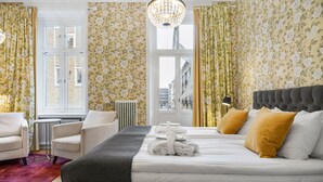 Une chambre d’hôtel avec un lit, deux fauteuils, un radiateur, une fenêtre avec des rideaux et un lustre.