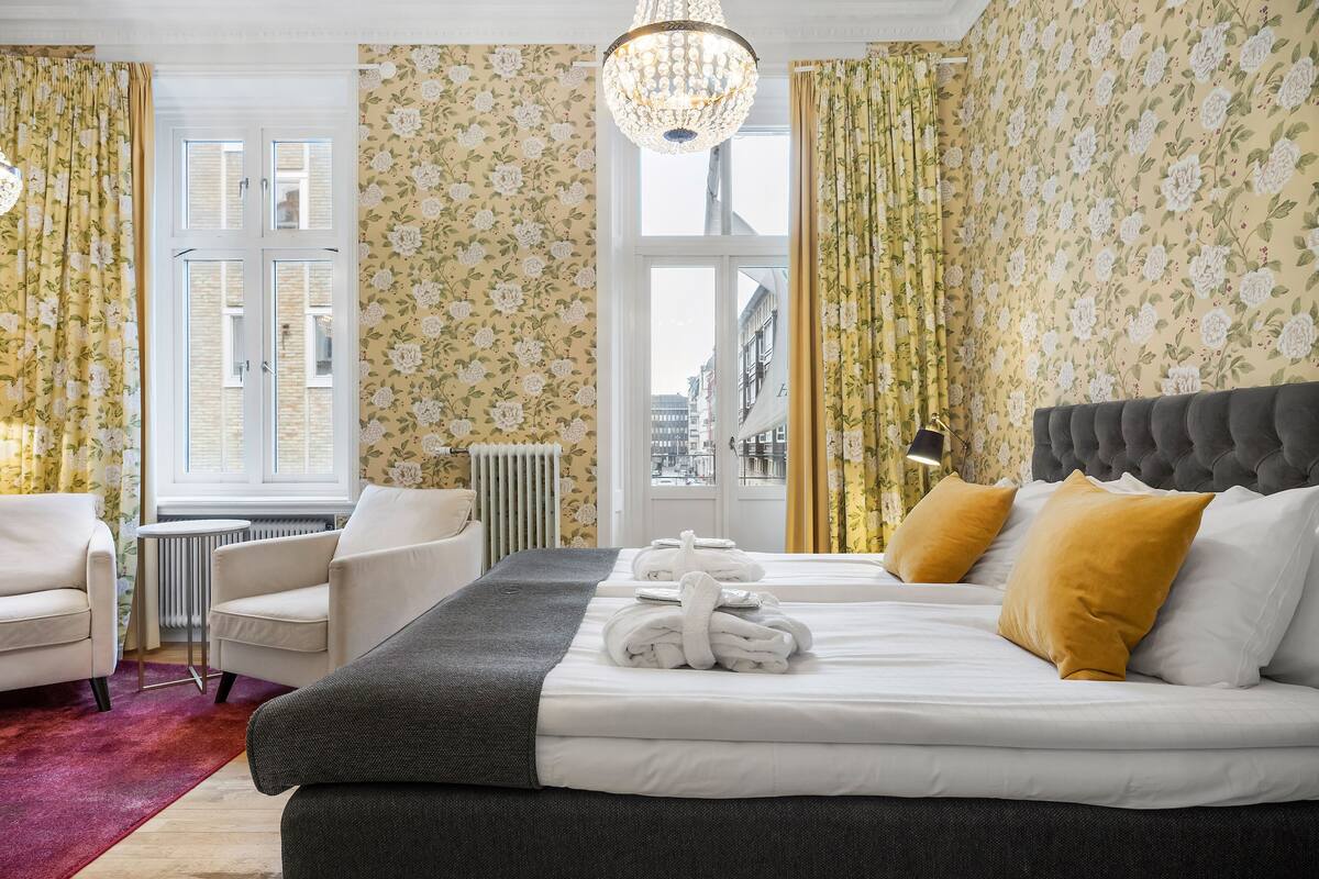 Quarto de hotel com cama, duas poltronas, radiador, janela com cortinas e um lustre.