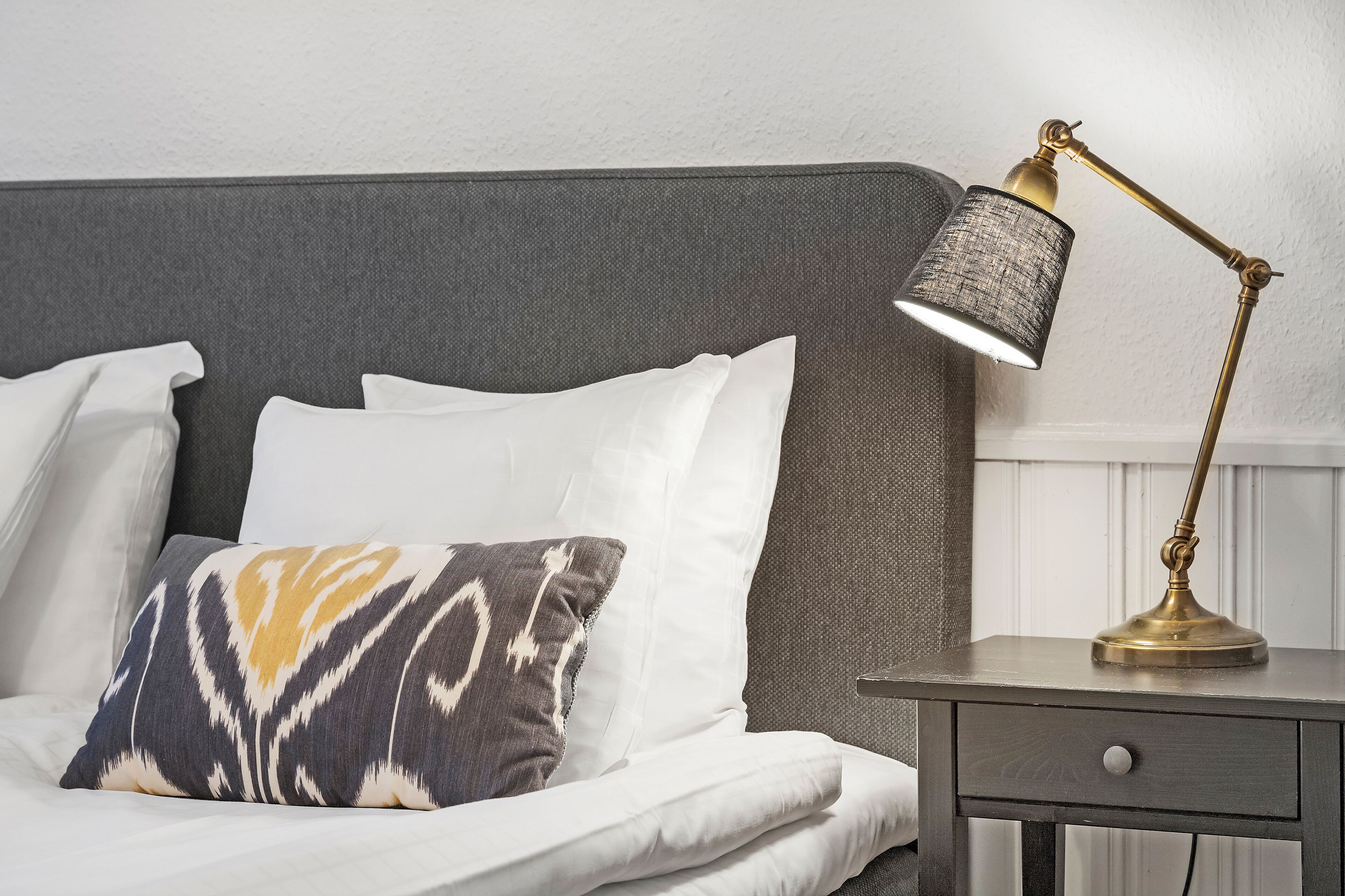 A neatly made bed with white linens and a patterned pillow, a bedside table with a lamp, and a gray headboard.