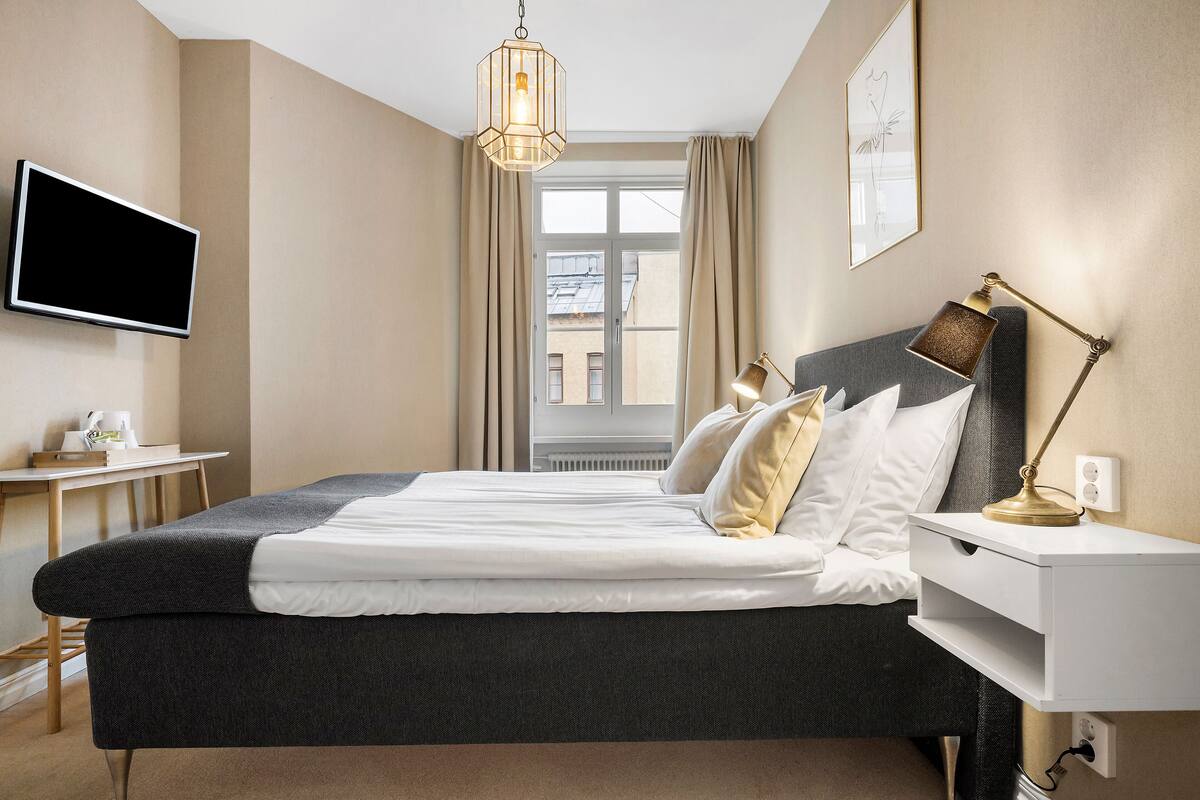 Quarto de hotel bem arrumado, com cama, mesa de cabeceira, abajur e televisão na parede.