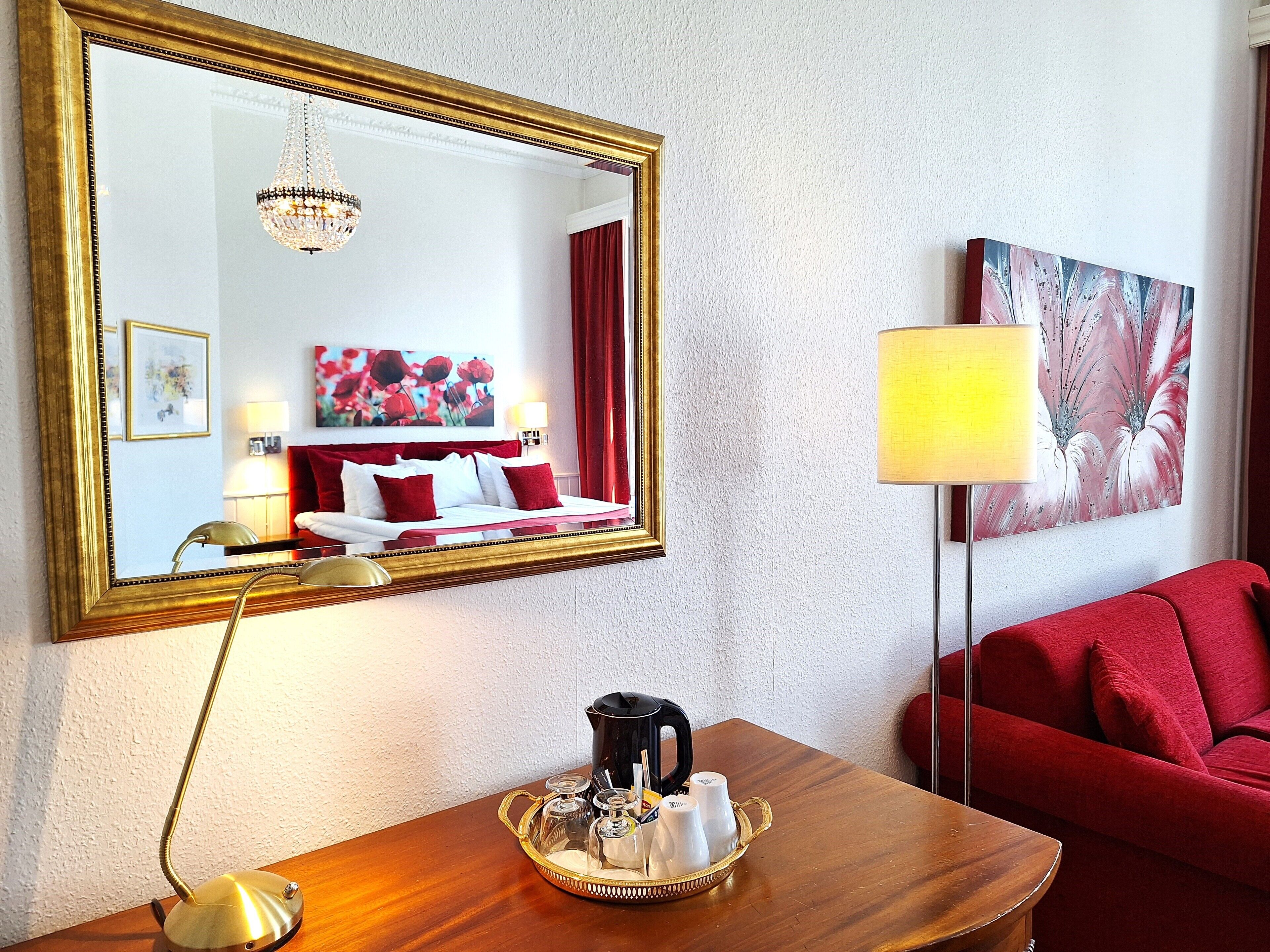 A hotel room with a large mirror, a bed with red bedding, a red sofa, a coffee table with a tray of cups, and a lamp.