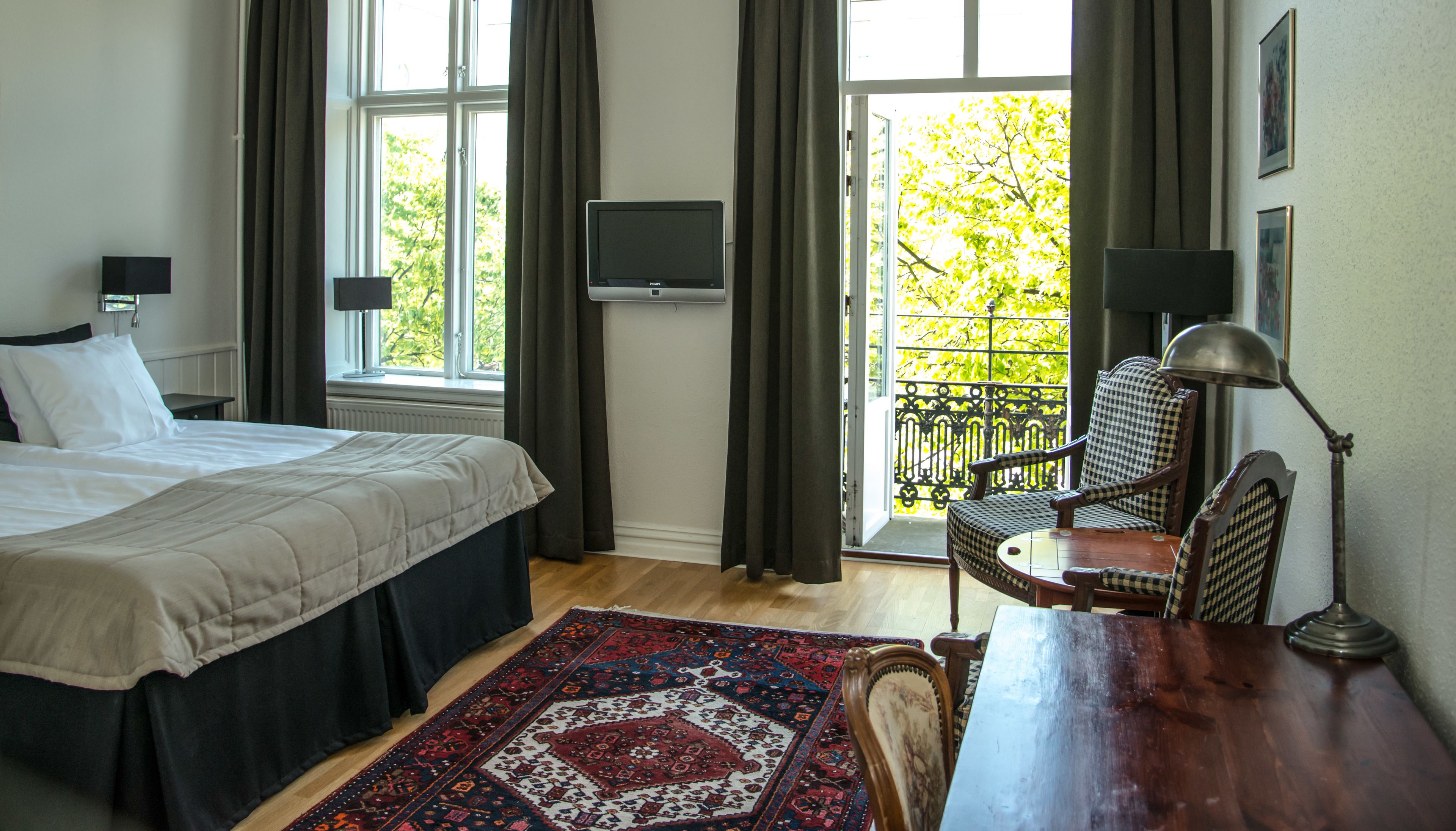 A bedroom with a bed, a desk, a chair, a TV, and a view of greenery through the window.