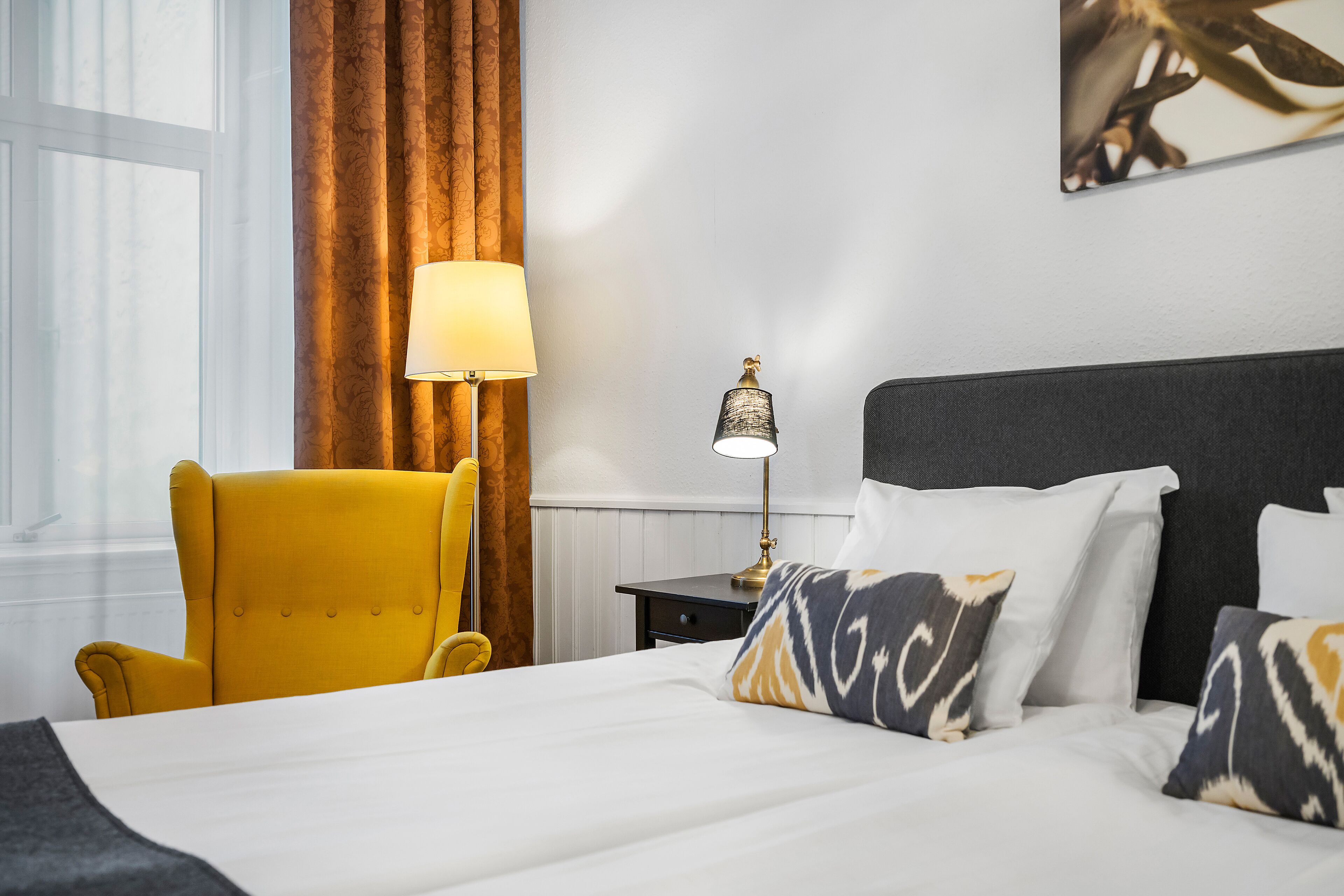 Una camera d'albergo con un letto, una poltrona gialla, un comodino con una lampada e un quadro appeso al muro.