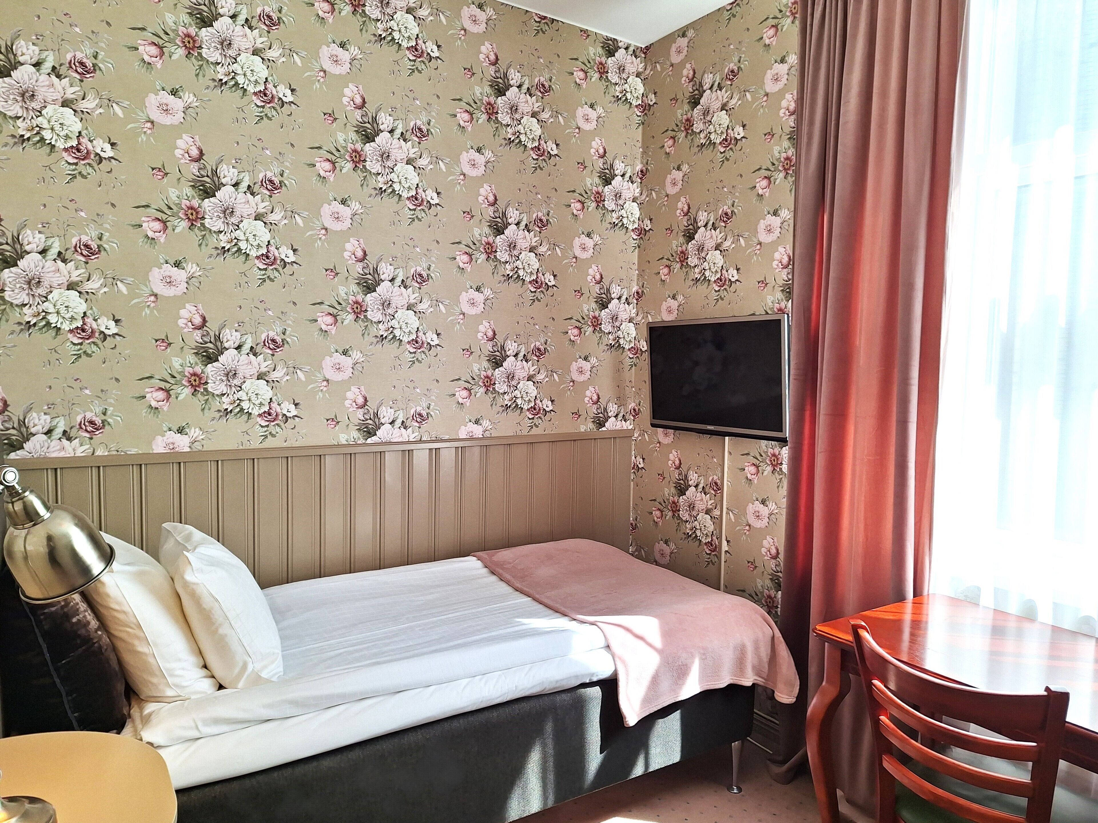 Ein Schlafzimmer mit Bett, Schreibtisch, Stuhl, Fernseher und blumigem Wandpapier.