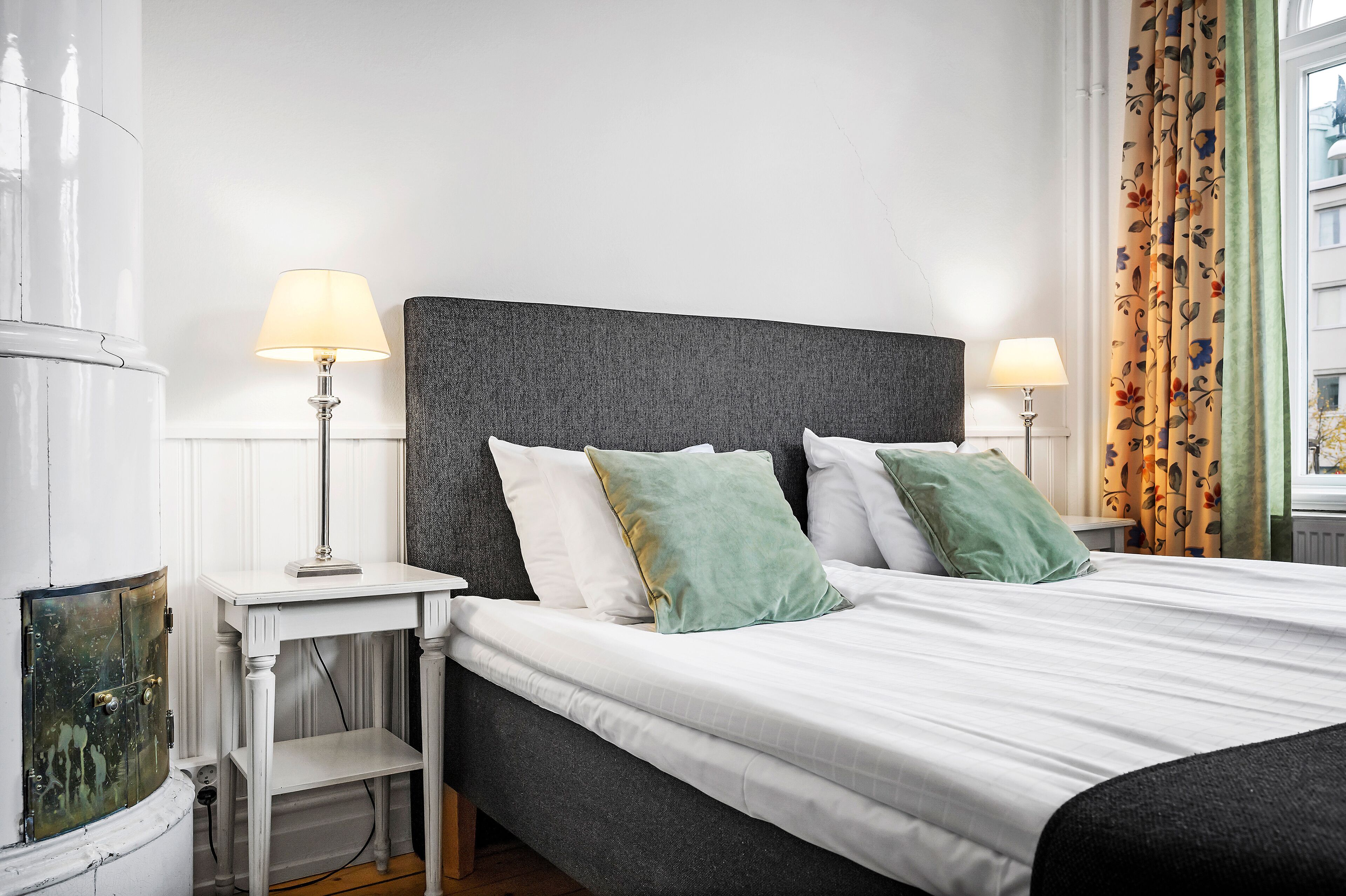 Una cama bien hecha con sábanas blancas y dos almohadas, una mesita de noche con una lámpara y una ventana con cortinas florales.