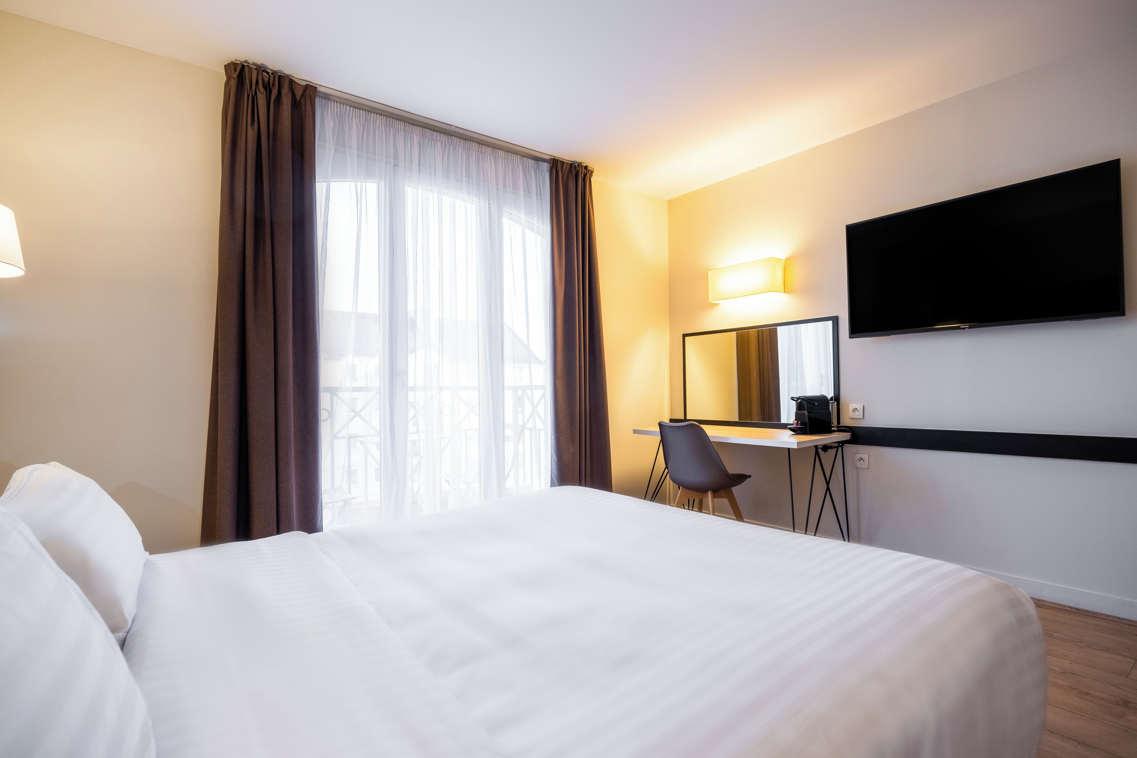 Habitación de hotel con cama, escritorio, silla, televisor de pantalla plana y ventana con cortinas.