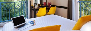 Habitación de hotel con cama, una computadora portátil, dos sillas amarillas, una mesita con cafetera y ventana que ofrece vistas a zonas verdes.