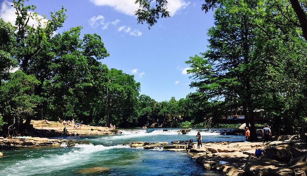 A river with rocks and trees, people relaxing by the water, and a clear blue sky.