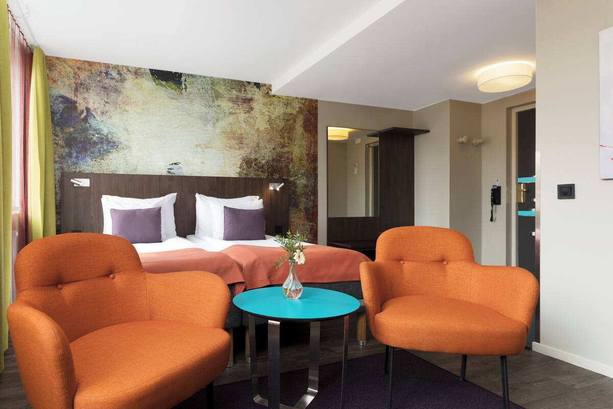 Ett hotellrum med en säng, två orangefärgade fåtöljer, ett litet runt bord och en färgstark, abstrakt väggmålning.
