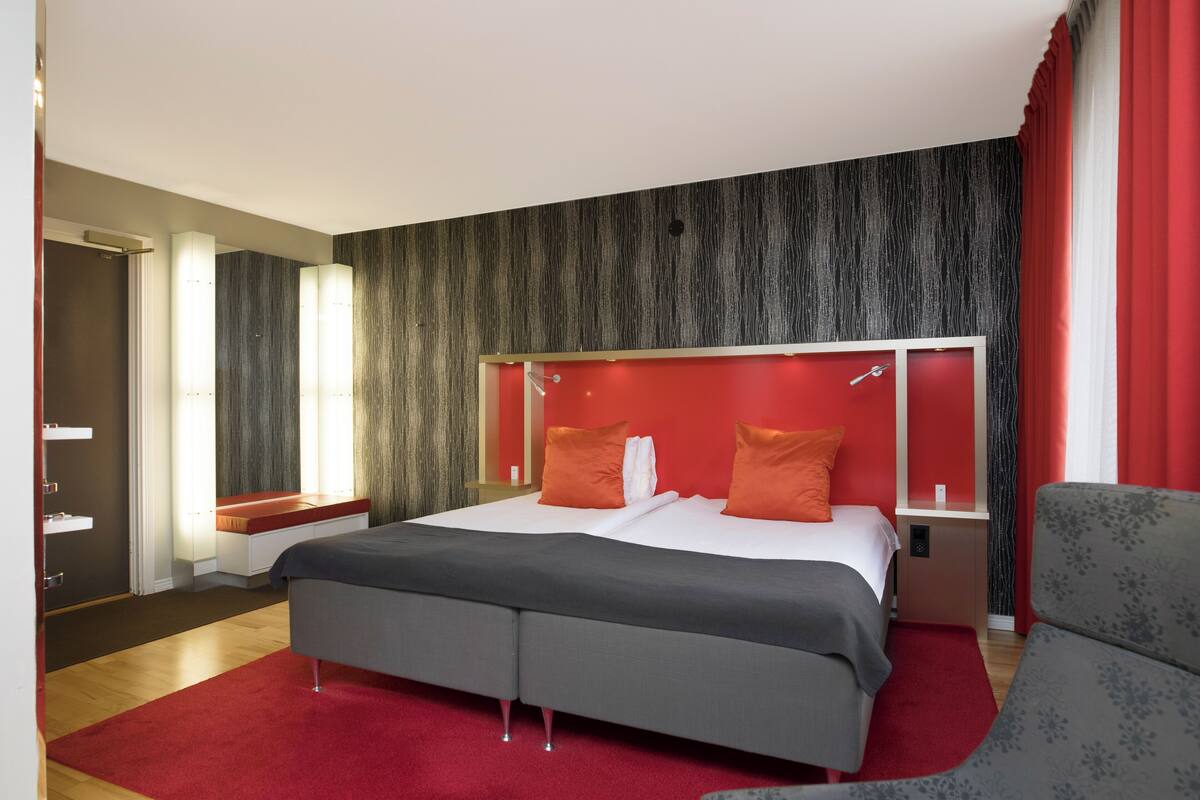 Ett modernt hotellrum med en stor säng, en röd accentvägg och en sittgrupp med en fåtölj.