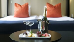A bed with white linens and orange pillows, a bottle of champagne in a metal bucket, and a tray with fruits and a glass of champagne. - ProfilHotels Savoy (Jonkoping)