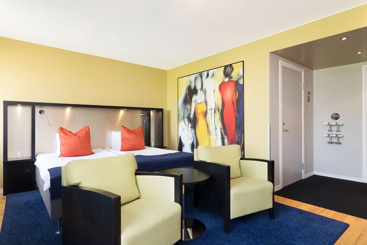Ett hotellrum med en säng, två stolar, ett bord, en tavla och en dörr.