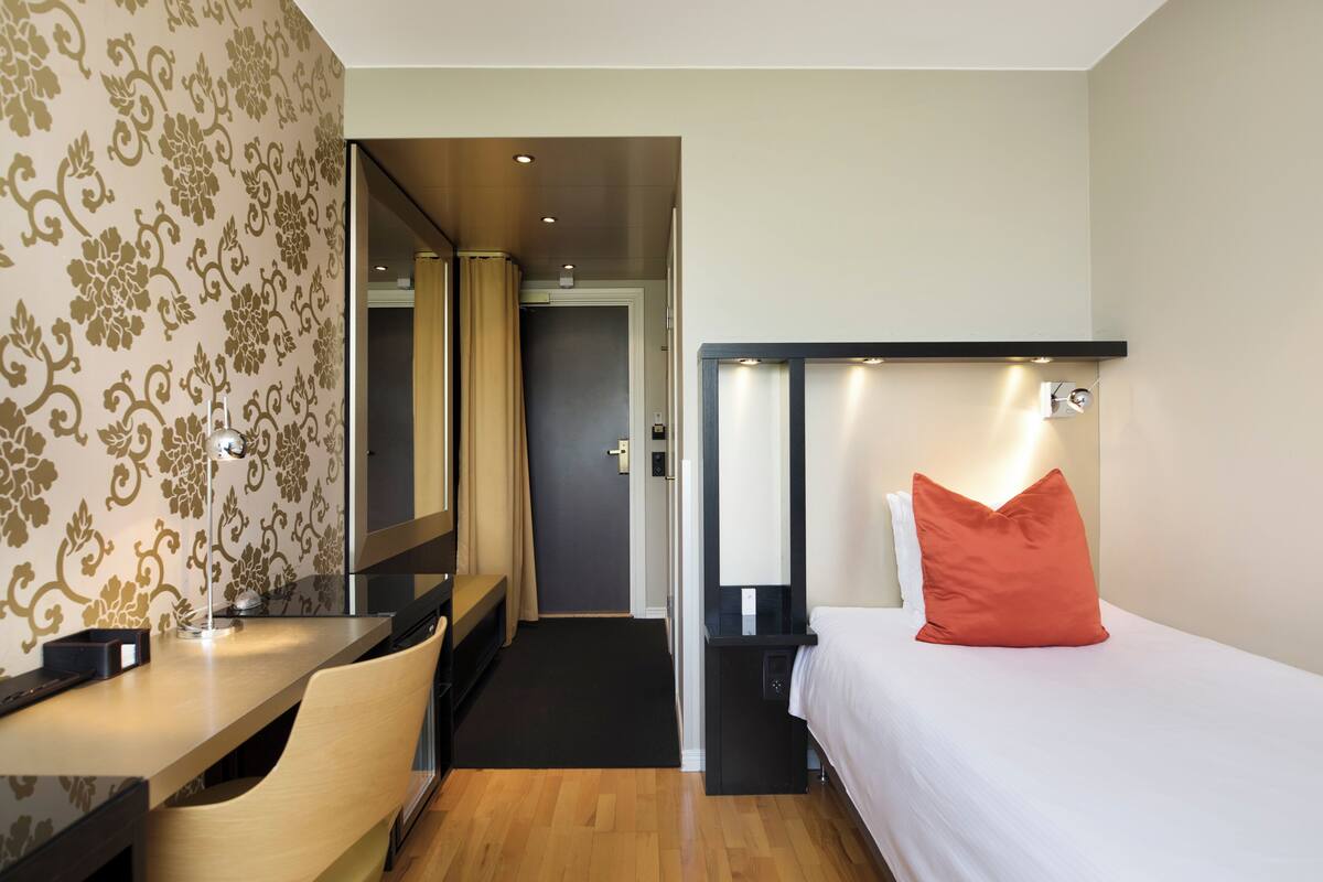 Ett hotellrum med ett skrivbord, en säng med en orangefärgad kudde, en spegel och en dörr som leder till ett annat rum.
