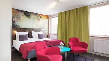 A hotel room with a bed, two red chairs, a small table, and a large abstract painting on the wall.