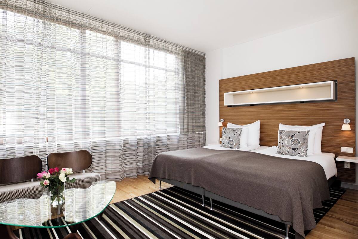 Ett modernt hotellrum med en stor säng, ett glasbord, en soffa och utsikt över omgivningarna genom tunna, genomskinliga gardiner.